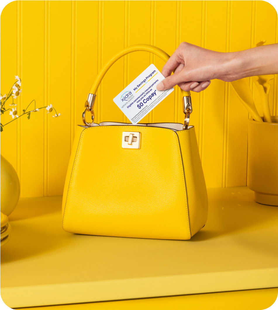 Hand holding a Xiidra Savings Program card above a yellow purse on a yellow counter